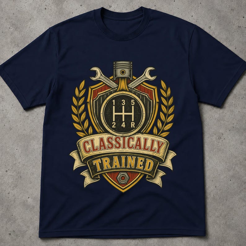 May include: A navy blue t-shirt featuring a graphic print of a manual transmission gear shift pattern, a piston, and two crossed wrenches inside a shield emblem. The design includes wheat stalks and a banner with the text "CLASSICALLY TRAINED".