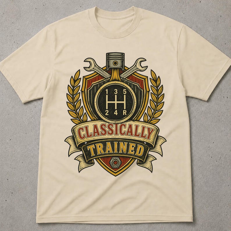 May include: A beige short-sleeved t-shirt featuring a graphic print of a shield, a piston, and two crossed wrenches. The design includes a manual transmission gear shift pattern and a banner with the text "CLASSICALLY TRAINED" in bold lettering.