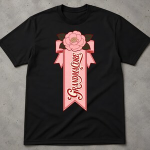 Pode incluir: Uma t-shirt preta de manga curta com uma impressão gráfica de uma flor cor-de-rosa sobre uma fita cor-de-rosa. A fita contém o texto "GRANDMACORE" numa fonte vermelha estilizada, com folhas verde-escuras a emoldurar a base da flor.