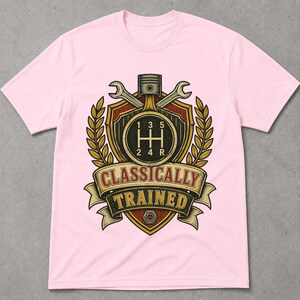 May include: A light pink t-shirt featuring a graphic of a car gear shift pattern inside a shield, flanked by two wrenches and wheat stalks. Above the shield is a piston. A banner below reads "CLASSICALLY TRAINED" with a metal nut at the bottom.