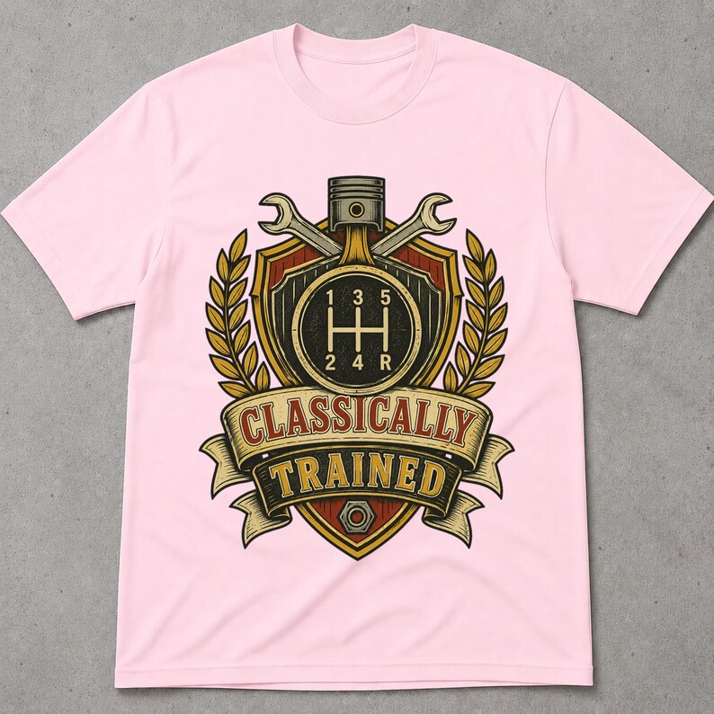 May include: A light pink t-shirt featuring a graphic of a car gear shift pattern inside a shield, flanked by two wrenches and wheat stalks. Above the shield is a piston. A banner below reads "CLASSICALLY TRAINED" with a metal nut at the bottom.