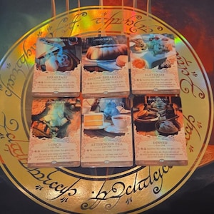 May include: Six rectangular game cards arranged in two rows of three, featuring illustrations of various meals labeled Breakfast, Second Breakfast, Elevenses, Lunch, Afternoon Tea, and Dinner, resting on a circular gold emblem with script text.