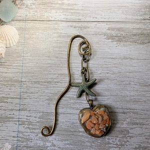 Puede incluir: Un marcapáginas de metal en tono bronce con un diseño de gancho curvo y un dije colgante. El dije consiste en una pequeña estrella de mar verde texturizada unida a un colgante en forma de corazón relleno de fragmentos de piedra naranja-marrón.