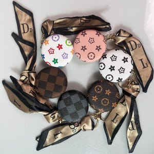 May include: A collection of round coin purses with various patterns, including stars, logos, and checkered designs. Each purse is attached to a gold-colored keychain and a tan and black patterned ribbon.