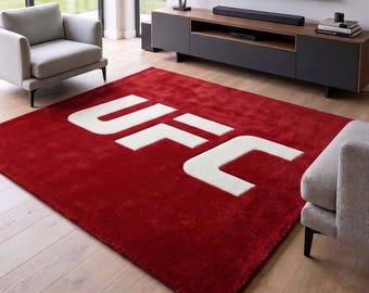 May include: A rectangular area rug featuring a deep red pile with the letters UFC displayed in large, bold, white block lettering across the center. The rug has a soft, textured surface and is positioned on a light-colored wooden floor.