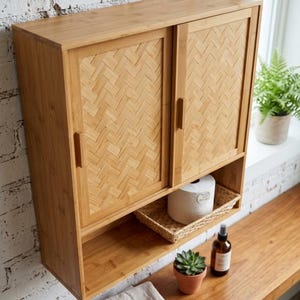 May include: A light brown bamboo wall-mounted cabinet with sliding doors and a woven pattern. The cabinet has a shelf below with a woven basket, a roll of toilet paper, a small potted plant, and a spray bottle. A folded towel is on the shelf.
