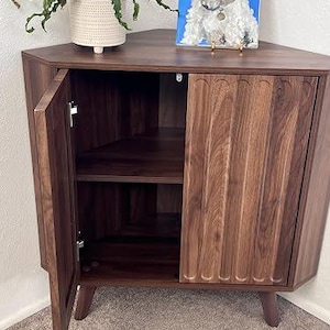 Pu&ograve; includere: Un mobile angolare in legno marrone scuro con piano triangolare, due ante con dettagli scanalati verticali e gambe affusolate. L'anta sinistra &egrave; aperta, rivelando un ripiano interno. Una pianta in vaso bianco e un dipinto di un cane sono appoggiati sopra.
