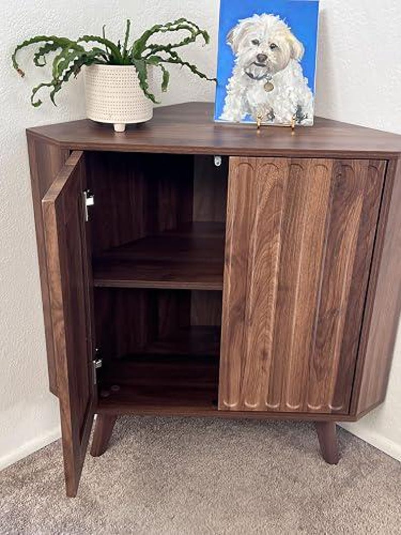 Pu&ograve; includere: Un mobile angolare in legno marrone scuro con piano triangolare, due ante con dettagli scanalati verticali e gambe affusolate. L'anta sinistra &egrave; aperta, rivelando un ripiano interno. Una pianta in vaso bianco e un dipinto di un cane sono appoggiati sopra.