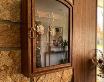 Può includere: Un mobiletto portachiavi in legno con porta a vetro, montato su un muro di pietra. Il mobiletto ha una finitura marrone scuro e un bordo decorativo. Diverse chiavi e una chiave dell'auto sono visibili all'interno.