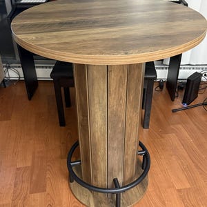 May include: A round pub table with a wood grain finish featuring a cylindrical pedestal base composed of vertical panels and a circular black metal footrest ring attached to the base, standing on a matching circular wood grain base plate.