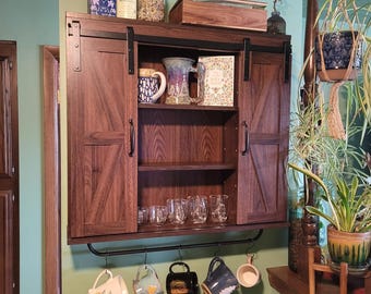 Può includere: Un mobile in legno scuro con ante scorrevoli, contenente tazze, bicchieri e oggetti decorativi. Sotto, una barra di metallo nero mostra tazze appese. Il mobile è montato su una parete verde, con piante sullo sfondo.