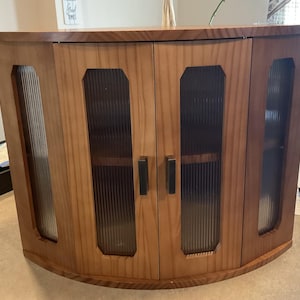 May include: A wooden cabinet with a curved front and four doors. The doors have ribbed glass panels and dark metal handles. The cabinet is made of light and dark brown wood.