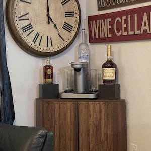 Pu&ograve; includere: Un mobile in legno scuro con ante a scanalature verticali, sormontato da una macchina per bevande argentata, due bicchieri trasparenti e bottiglie di Grey Goose e Hennessy. Sopra il mobile &egrave; appeso un grande orologio da parete con numeri romani.