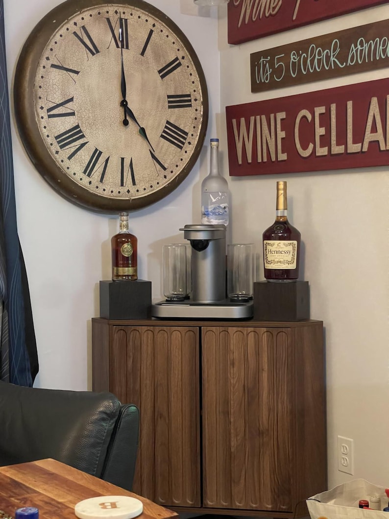 Pu&ograve; includere: Un mobile in legno scuro con ante a scanalature verticali, sormontato da una macchina per bevande argentata, due bicchieri trasparenti e bottiglie di Grey Goose e Hennessy. Sopra il mobile &egrave; appeso un grande orologio da parete con numeri romani.