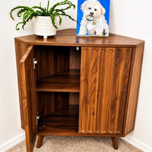 May include: A dark wood corner cabinet with a triangular top and tapered legs. The cabinet features one open door revealing an interior shelf and one closed door with vertical fluted detailing. A white potted plant and a dog portrait sit on top.