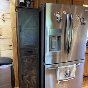 May include: A dark brown wooden cabinet with a drawer and a door, next to a stainless steel refrigerator. The cabinet has a rustic design with a diagonal pattern on the door. A black container sits on top of the cabinet.