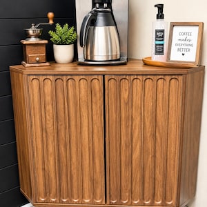 Pu&ograve; includere: Un mobile angolare in legno marrone con due ante a scanalature verticali e gambe affusolate. Sopra si trovano una macchina per il caff&egrave; in acciaio inossidabile, un macinacaff&egrave; manuale, una piccola pianta, un dispenser di sapone e una cornice con la scritta COFFEE makes EVERYTHING BETTER.