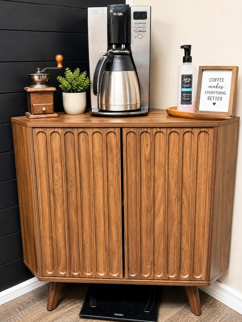 Pu&ograve; includere: Un mobile angolare in legno marrone con due ante a scanalature verticali e gambe affusolate. Sopra si trovano una macchina per il caff&egrave; in acciaio inossidabile, un macinacaff&egrave; manuale, una piccola pianta, un dispenser di sapone e una cornice con la scritta COFFEE makes EVERYTHING BETTER.