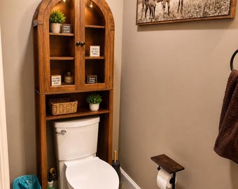 Può includere: Un mobiletto alto in legno ad arco da posizionare sopra il WC, dotato di ante in vetro, illuminazione interna e ripiani a giorno. Il mobile contiene piccole piante in vaso, un cestino intrecciato e targhette decorative sopra un WC bianco.