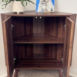 Pu&ograve; includere: Un mobile ad angolo in legno marrone scuro con due ante aperte, un ripiano centrale e quattro gambe affusolate. La superficie del mobile sostiene un vaso bianco testurizzato con una pianta verde e un dipinto su tela blu di un cane bianco.