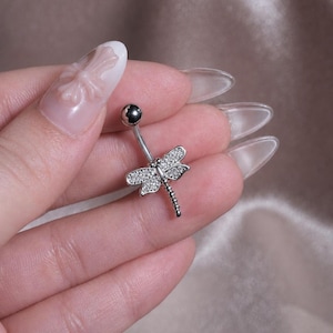 May include: A silver-toned navel ring featuring a dragonfly charm. The dragonfly wings are encrusted with small, clear, sparkling stones, and the body is a slender metal segment. The barbell has a smooth, round, dark-colored ball at the top end.