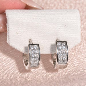 May include: A pair of silver-toned hoop earrings featuring a double row of small, clear, round-cut stones embedded along the front surface. The earrings are secured to a rectangular piece of white fabric, showcasing a polished metallic finish.
