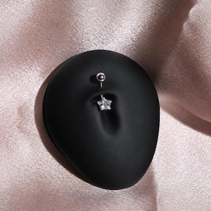 May include: A silver-toned belly button ring featuring a small, clear, star-shaped crystal charm at the bottom and a smooth, round silver-toned ball at the top, designed for navel piercing jewelry, displayed on a matte black surface.