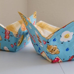 May include: Two square fabric bowl cozies. The exterior is light blue with a repeating pattern of smiling breakfast foods like waffles, eggs, and bacon. The interior is yellow and white checkered fabric. The cozies are folded into a bowl shape.