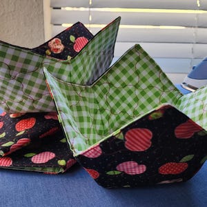 May include: Three fabric bowls with a green and white gingham pattern and a black fabric with red and white apple print. The bowls are folded and stacked on a blue surface.
