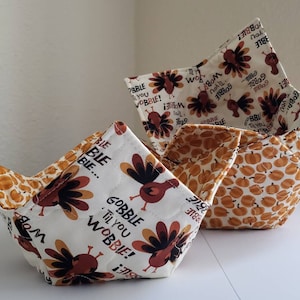 May include: Two fabric bowls with a Thanksgiving theme. One bowl is white with a brown turkey print and the text "Gobble Til You Wobble!" The other bowl is white with a brown pumpkin print.