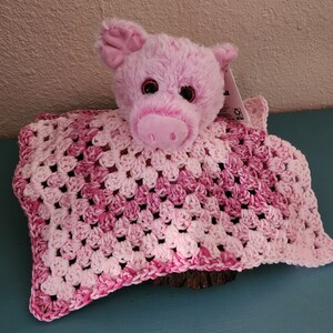 May include: A pink plush pig with large eyes sits atop a handmade crochet blanket. The blanket features a granny square design in shades of pink and white. The pig has a soft, fuzzy texture.