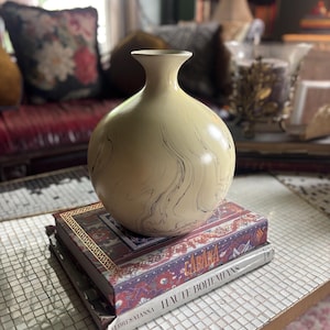 May include: A round, cream-colored ceramic vase with a narrow neck and a subtle black marbled pattern. The vase is resting on top of two stacked hardcover books, one titled Cabana and the other titled Haute Bohemians by Flores-Vianna.