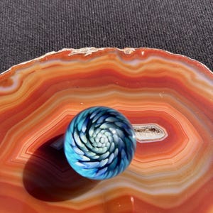May include: A blue and white glass marble with a spiral design, resting on an orange and white agate slice. The marble's interior features a swirling pattern of blue and white, creating a captivating visual effect.