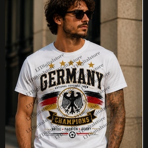 Könnte beinhalten: Ein weißes Kurzarm-T-Shirt mit einem Grafikdruck und dem Text GERMANY NATIONAL FOOTBALL TEAM CHAMPIONS PRIDE PASSION GLORY. Das Design zeigt einen schwarzen Adler, die deutsche Flagge, fünf goldene Sterne und einen Fußball.