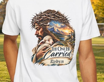 May include: A white t-shirt featuring a graphic of a figure wearing a crown of thorns and holding a lamb. The design includes a landscape with mountains and a sunset, with the text "THE ONE HE Carried" and the name "Robyn" printed below.