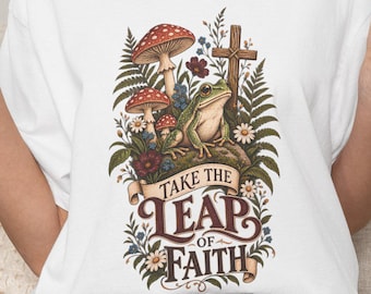 May include: A white t-shirt featuring a graphic print of a green frog sitting on a rock surrounded by mushrooms, ferns, and a wooden cross. Below the illustration, a banner contains the text "TAKE THE LEAP OF FAITH" in dark brown lettering.
