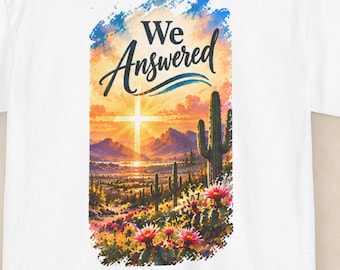 May include: A white t-shirt featuring a rectangular graphic print on the back. The design depicts a desert landscape with cacti, blooming flowers, mountains, and a glowing cross under a sunset sky, with the text "We Answered" in black script.