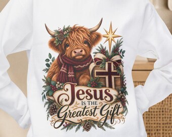 May include: A white long-sleeve sweatshirt featuring a graphic of a Highland cow wearing a scarf, surrounded by pine branches, a star, and a cross. The text reads Jesus is the Greatest Gift in brown script and block lettering.