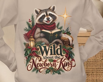 May include: A beige long-sleeve sweatshirt featuring a graphic of a raccoon wearing a red scarf, reading a book, surrounded by pine branches, a lantern, and a star. The text reads, Wild About the Newborn King, in decorative lettering.