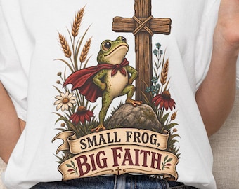 May include: A white t-shirt featuring a graphic of a green frog wearing a red cape, standing on a rock next to a wooden cross. The design includes wheat, flowers, and a banner with the text "SMALL FROG, BIG FAITH" in brown lettering.