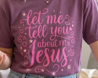 May include: A short-sleeved, plum-colored crew neck t-shirt featuring the text "let me tell you about my Jesus" printed in a bright pink, cursive font. The design is accented with small stars, swirls, and heart graphics surrounding the lettering.