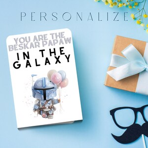 Może przedstawiać: Kartka z pozdrowieniami z napisem "YOU ARE THE BESKAR PAPAW IN THE GALAXY" i ilustracją postaci trzymającej balony. Widoczne jest r&oacute;wnież zapakowane pudełko prezentowe z jasnoniebieską wstążką oraz rekwizyt z wąsami i okularami.