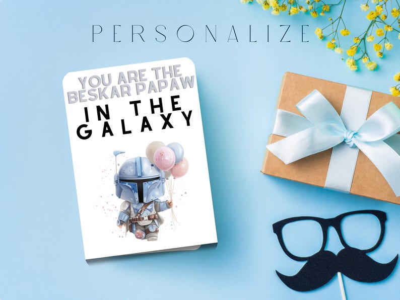 Może przedstawiać: Kartka z pozdrowieniami z napisem "YOU ARE THE BESKAR PAPAW IN THE GALAXY" i ilustracją postaci trzymającej balony. Widoczne jest r&oacute;wnież zapakowane pudełko prezentowe z jasnoniebieską wstążką oraz rekwizyt z wąsami i okularami.