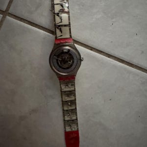 May include: A silver-toned wristwatch with a transparent band featuring black and white abstract designs. The watch has a red accent on the band and a circular face with visible internal mechanisms. The watch is lying on a tiled surface.