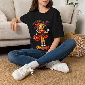 May include: A black t-shirt featuring a graphic of a cartoon bee dressed as a witch, wearing a hat and holding a jack-o'-lantern bucket. The character has curly hair, wings, and striped socks. Below the graphic, the text reads Bee Spooky.