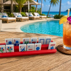 May include: A set of white rectangular tiles with colorful printed symbols and numbers arranged on a red plastic rack. The tiles feature various designs including flowers, birds, dots, and a Joker card, all displayed on a wooden surface next to a glass.