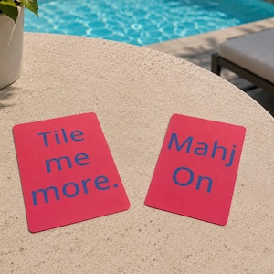 May include: Two rectangular pink cards with blue text on a beige table. One card reads "Tile me more." The other card reads "Mahj On." The cards are likely for a tile-based game.