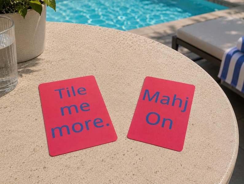 May include: Two rectangular pink cards with blue text on a beige table. One card reads "Tile me more." The other card reads "Mahj On." The cards are likely for a tile-based game.