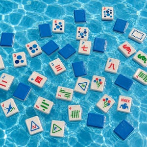 May include: A collection of Mahjong tiles floating in blue water. The tiles are white with various symbols and numbers in red, green, and blue, and are interspersed with solid blue tiles. The scene is brightly lit, suggesting a sunny day.
