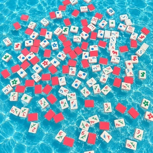 May include: A collection of rectangular Mahjong tiles floating in clear blue water. The tiles feature white faces with various colorful symbols, numbers, and Chinese characters, while the backs of the tiles are a solid, vibrant red color.